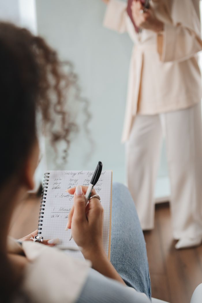 Close-up of a student jotting down notes during a class with a teacher in the background.